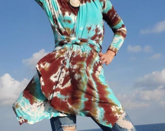 May include: A long-sleeved tie-dye top in shades of turquoise, brown, and white. The top is paired with distressed blue jeans. A large pendant necklace is visible. The background features a blue sky and ocean.