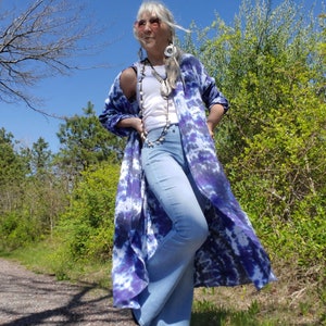 Könnte beinhalten: Eine Frau trägt ein weißes Tanktop, blaue Jeans und ein langes, fließendes Batik-Shirt in Blautönen und Lila. Sie steht auf einem Weg in einem Waldgebiet.