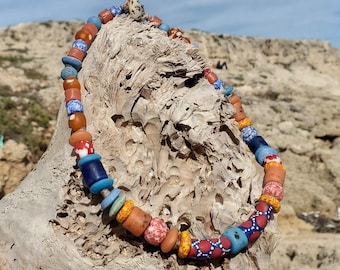 Puede incluir: Un collar de cuentas de colores con una variedad de formas y tamaños. Las cuentas son en tonos de azul, rojo, naranja y amarillo. El collar se exhibe sobre un trozo de madera flotante contra un paisaje rocoso y un cielo azul.