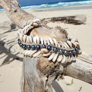 Puede incluir: Un collar de conchas blancas con una hebra de perlas azul oscuro. El collar está sobre un trozo de madera a la deriva sobre un fondo de playa.