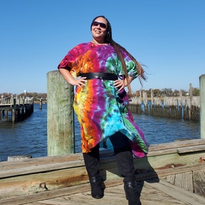 May include: A woman wearing a colorful tie-dye dress with a black belt and black boots. She is standing on a wooden dock with a body of water in the background.