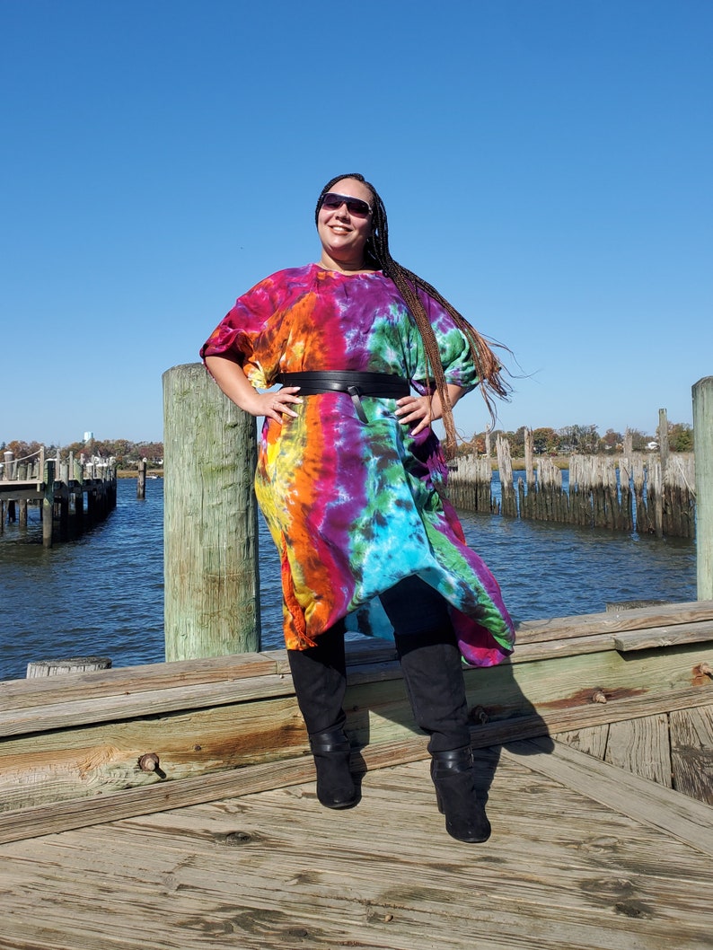 May include: A woman wearing a colorful tie-dye dress with a black belt and black boots. She is standing on a wooden dock with a body of water in the background.