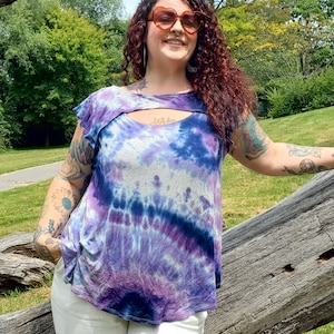 Puede incluir: Una blusa teñida con tintes de morado, azul y blanco. La blusa tiene un escote de ojo de cerradura y mangas cortas. La persona lleva pantalones blancos y gafas de sol en forma de corazón. El fondo es un parque con árboles y hierba.