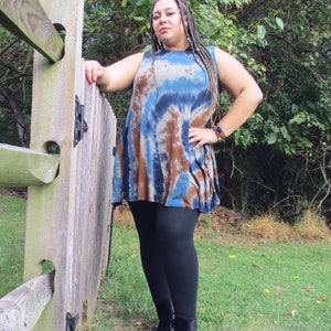 May include: A sleeveless tie-dye tunic in shades of blue and brown, paired with black leggings and black ankle boots. The model is leaning against a wooden fence in a grassy outdoor setting.