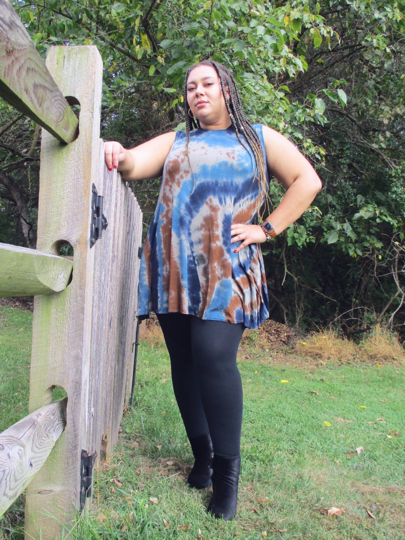 May include: A sleeveless tie-dye tunic in shades of blue and brown, paired with black leggings and black ankle boots. The model is leaning against a wooden fence in a grassy outdoor setting.
