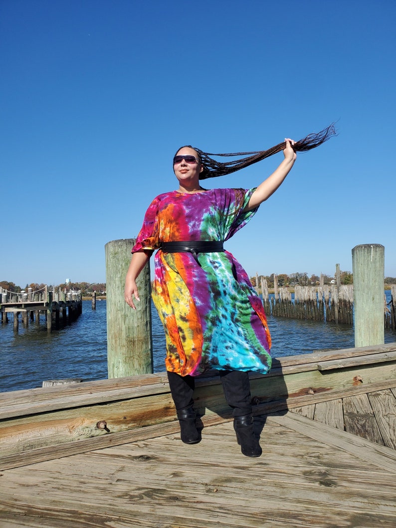 May include: A person wearing a colorful tie-dye maxi dress with a black belt and black boots. The person is standing on a wooden dock with a body of water in the background.