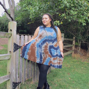 May include: A sleeveless tie-dye dress in shades of blue and brown. The dress has a flared silhouette and is paired with black leggings and ankle boots. The background features a wooden fence and lush greenery.