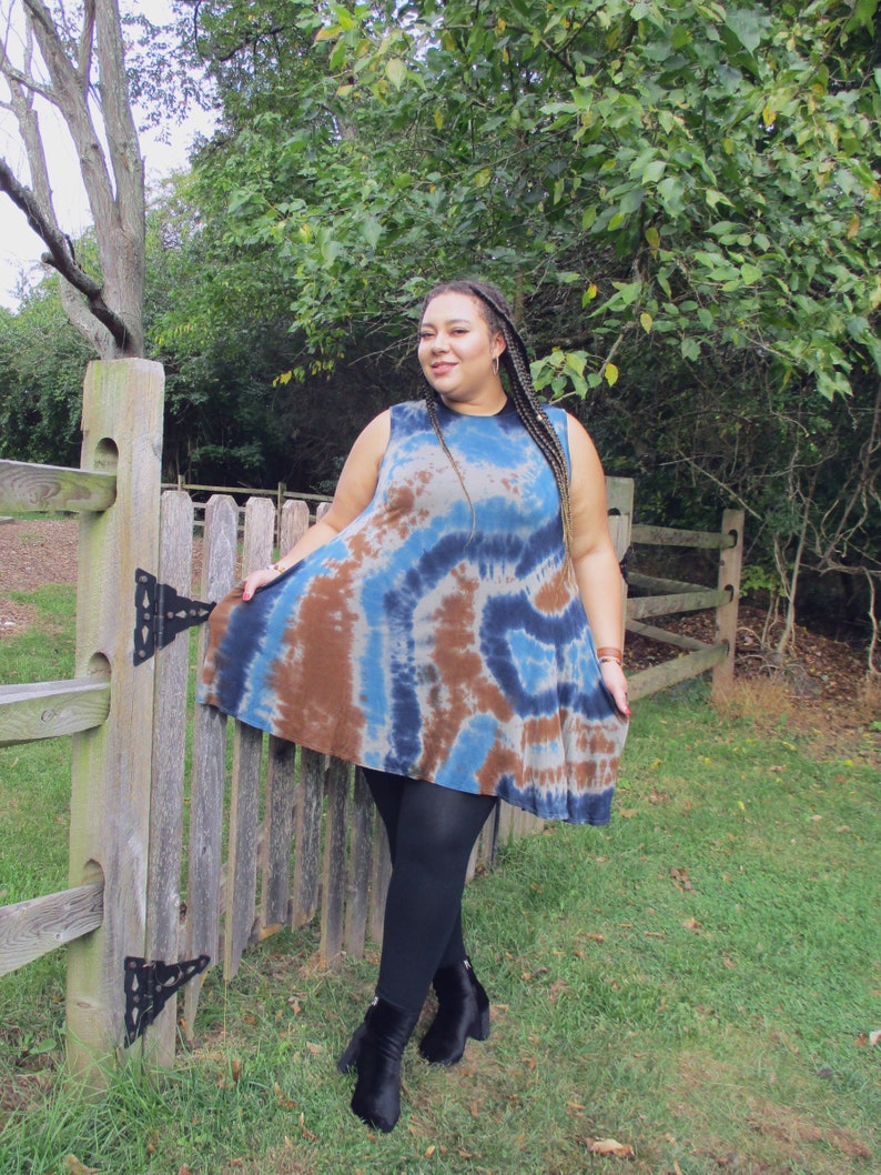 May include: A sleeveless tie-dye dress in shades of blue and brown. The dress has a flared silhouette and is paired with black leggings and ankle boots. The background features a wooden fence and lush greenery.