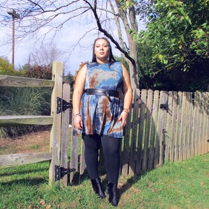 May include: A sleeveless tie-dye dress in shades of blue, brown, and white, cinched at the waist with a black belt. The dress is paired with black leggings and ankle boots. The background includes a wooden fence and green foliage.