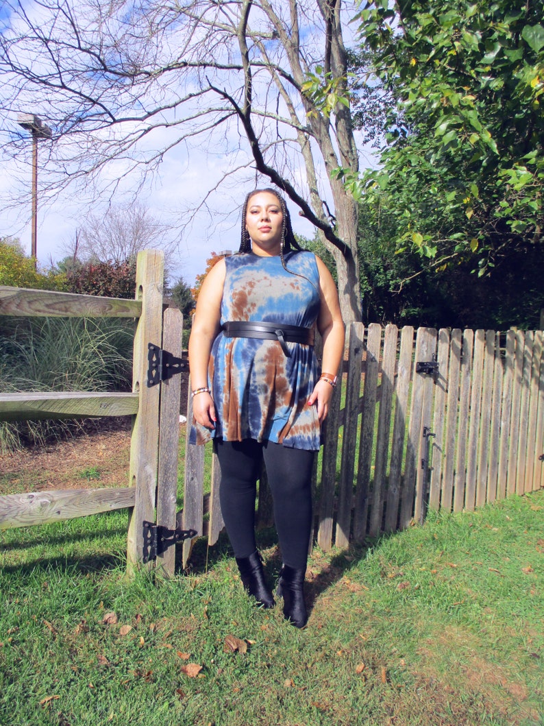May include: A sleeveless tie-dye dress in shades of blue, brown, and white, cinched at the waist with a black belt. The dress is paired with black leggings and ankle boots. The background includes a wooden fence and green foliage.