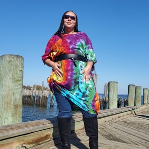 May include: A woman wearing a colorful tie-dye maxi dress with a black belt and black knee-high boots. She is standing on a wooden dock.