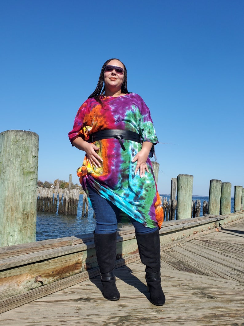 May include: A woman wearing a colorful tie-dye maxi dress with a black belt and black knee-high boots. She is standing on a wooden dock.