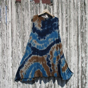 May include: A sleeveless, tie-dye tunic in shades of blue, brown, and gray hangs on a decorative hanger against a weathered white fence. The tunic features a flowing, A-line silhouette and a unique, swirling pattern, perfect for casual wear.