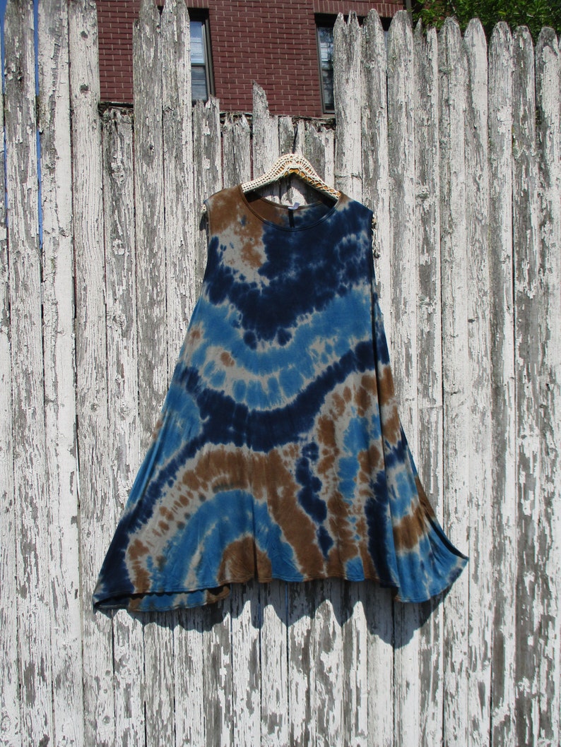 May include: A sleeveless, tie-dye tunic in shades of blue, brown, and gray hangs on a decorative hanger against a weathered white fence. The tunic features a flowing, A-line silhouette and a unique, swirling pattern, perfect for casual wear.