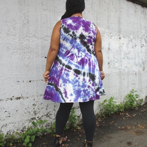 May include: A sleeveless tie-dye dress in shades of purple, blue, and black. The dress has a flared skirt and is paired with black leggings and sandals. The model is standing in front of a white wall.