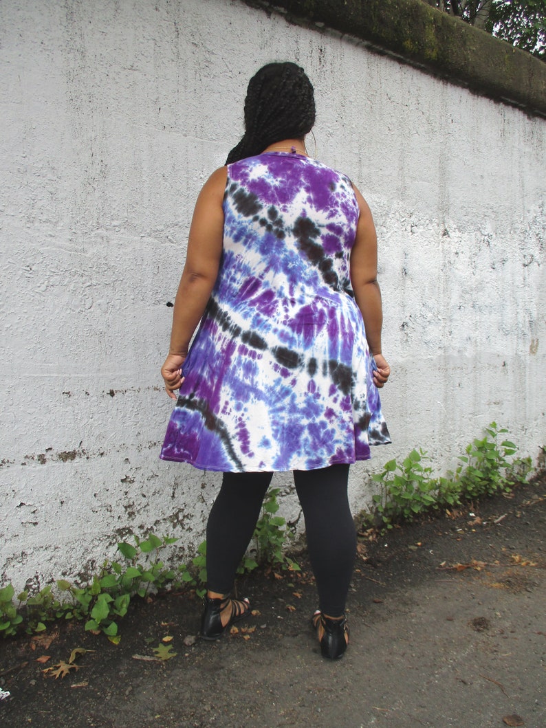 May include: A sleeveless tie-dye dress in shades of purple, blue, and black. The dress has a flared skirt and is paired with black leggings and sandals. The model is standing in front of a white wall.