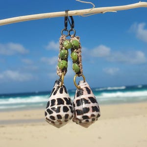 Puede incluir: Un par de pendientes con un colgante de concha de mar blanco y negro y cuentas verdes que cuelgan de un cordón marrón.