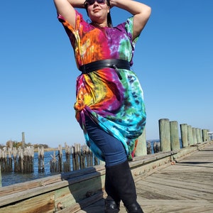 May include: A person wearing a colorful tie-dye maxi dress with a black belt and black boots. The person is standing on a wooden pier with water and pilings in the background.