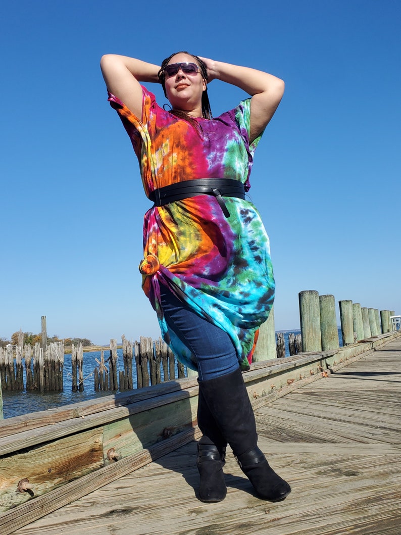 May include: A person wearing a colorful tie-dye maxi dress with a black belt and black boots. The person is standing on a wooden pier with water and pilings in the background.