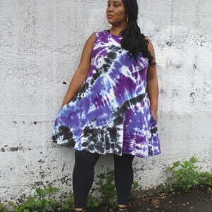 May include: A sleeveless tie-dye dress in shades of purple, black, and white. The dress has a flared silhouette and is paired with black leggings and black sandals. The model is standing in front of a white wall.