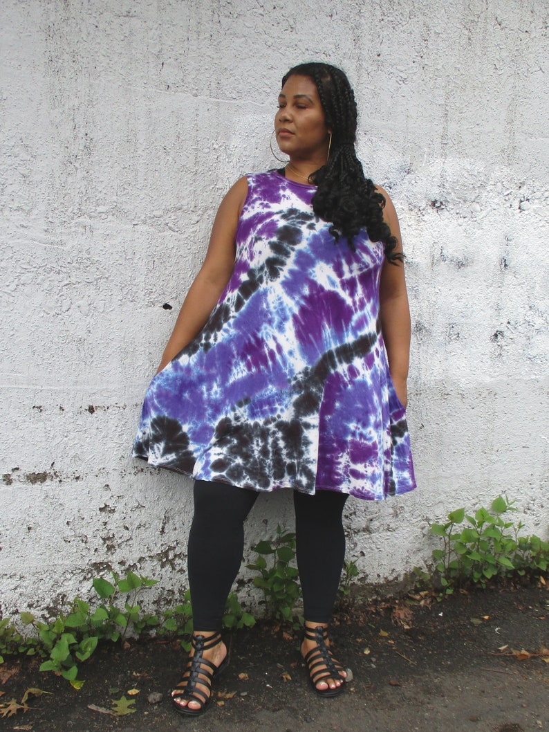 May include: A sleeveless tie-dye dress in shades of purple, black, and white. The dress has a flared silhouette and is paired with black leggings and black sandals. The model is standing in front of a white wall.