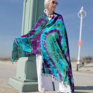 Può includere: Una donna indossa pantaloni bianchi e una grande sciarpa tie-dye colorata con sfumature di viola, blu e verde. La sciarpa ha un bordo a frangia ed è drappeggiata sulle sue spalle.