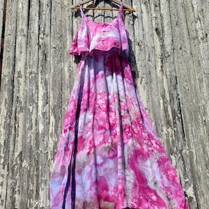 Puede incluir: Un vestido maxi de color rosa y morado con estampado tie-dye, con un top con volantes y tirantes finos. El vestido está colgado en una percha de madera contra una valla de madera desgastada.