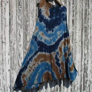 May include: A sleeveless tie-dye top in shades of blue, brown, and gray hangs on a white crocheted hanger. The fabric features a circular pattern of colors. The top has a flowing, asymmetrical design.