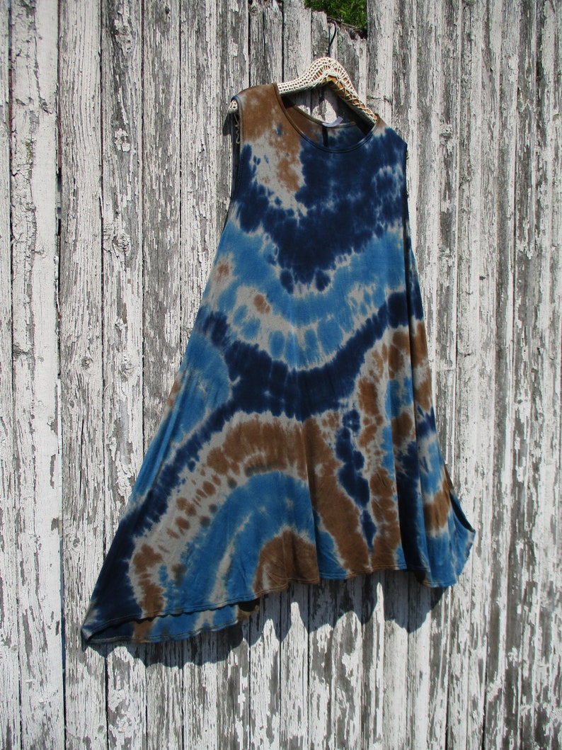 May include: A sleeveless tie-dye top in shades of blue, brown, and gray hangs on a white crocheted hanger. The fabric features a circular pattern of colors. The top has a flowing, asymmetrical design.