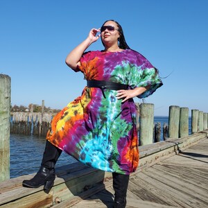 May include: A woman wearing a colorful tie-dye maxi dress with a black belt and black boots. She is standing on a wooden dock with a body of water in the background.