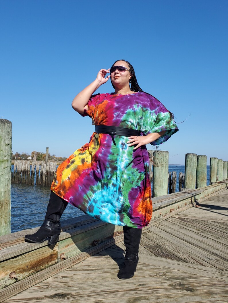 May include: A woman wearing a colorful tie-dye maxi dress with a black belt and black boots. She is standing on a wooden dock with a body of water in the background.