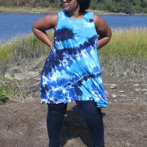 May include: A sleeveless, tie-dye tunic in shades of blue and navy. The tunic is paired with dark wash jeans and brown sandals. The background features a body of water and a clear blue sky.