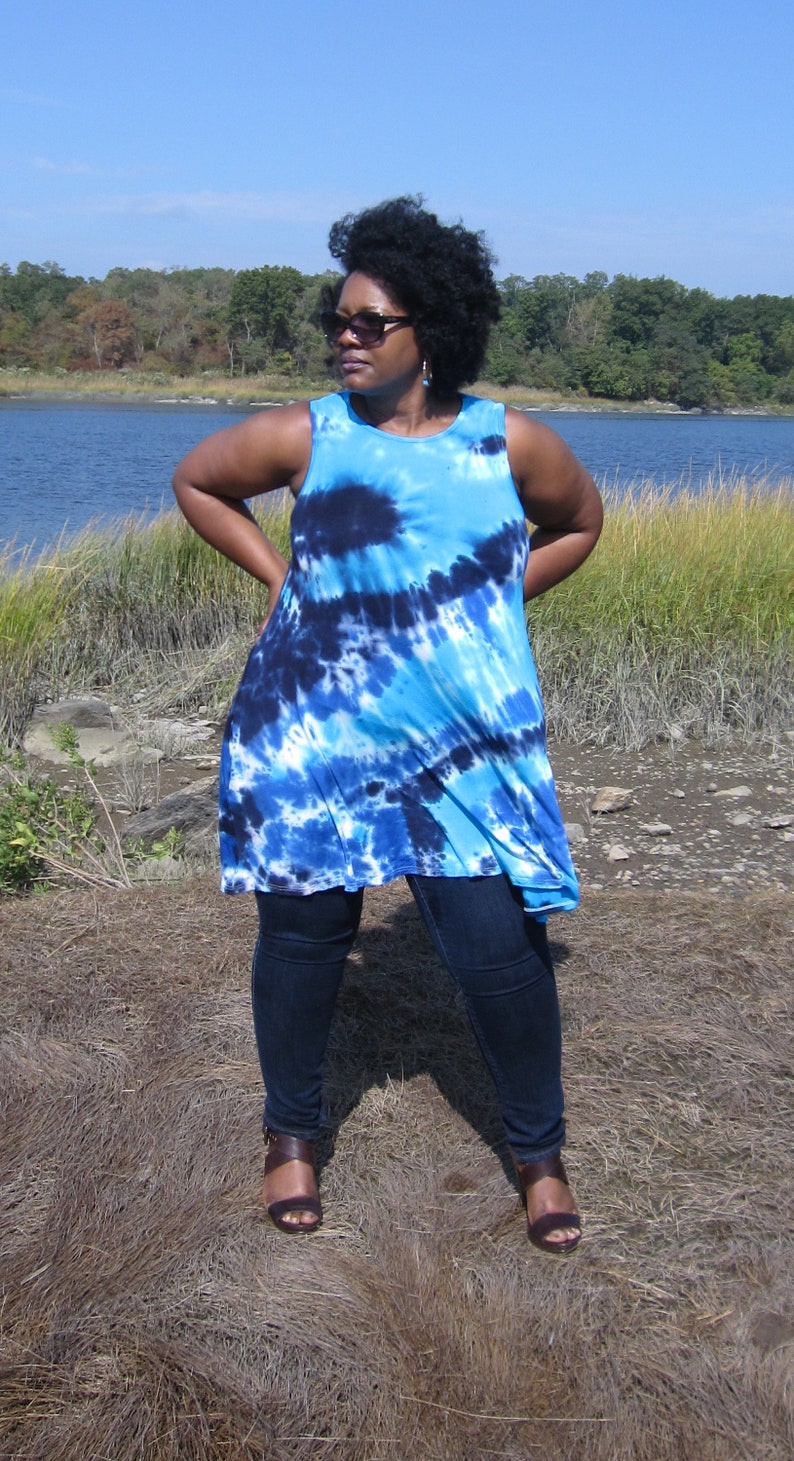 May include: A sleeveless, tie-dye tunic in shades of blue and navy. The tunic is paired with dark wash jeans and brown sandals. The background features a body of water and a clear blue sky.