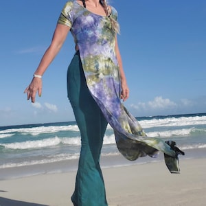 May include: A woman wearing a long, flowing tie-dye top with a side slit and a pair of dark green bell-bottom pants. She is standing on a sandy beach with blue water and white waves in the background.