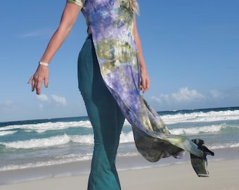 May include: A woman wearing a long, flowing tie-dye top with a side slit and a pair of dark green flared trousers. She is standing on a sandy beach with blue water and white waves in the background.