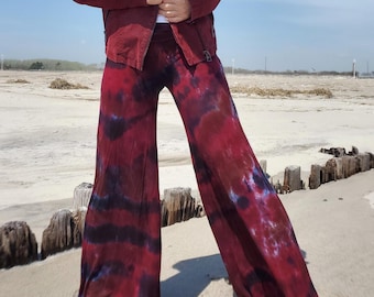 May include: A burgundy jacket and tie-dye pants ensemble. The jacket has a unique collar and a front zipper. The pants are wide-legged with a red and purple tie-dye pattern. The outfit is worn on a sandy beach.