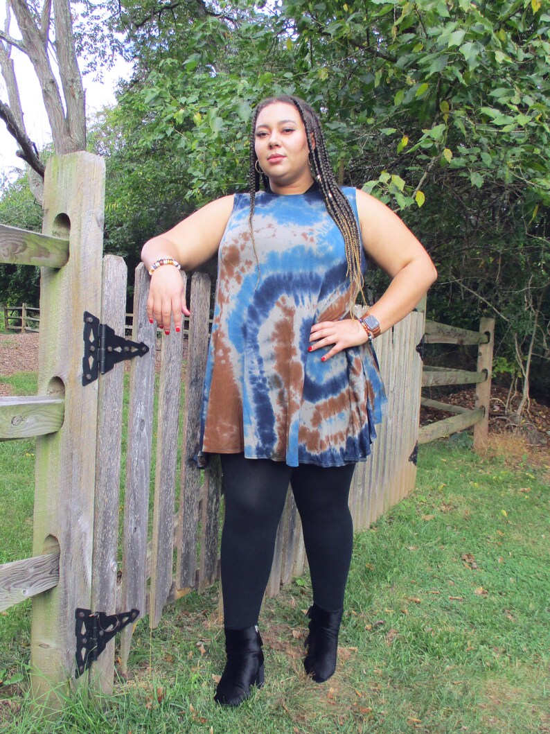 May include: A sleeveless tie-dye tunic in shades of blue, brown, and white. The tunic is paired with black leggings and black ankle boots. The woman is leaning against a wooden fence in an outdoor setting.