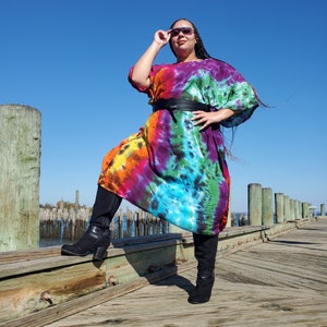May include: A woman wearing a colorful tie-dye maxi dress with a black belt and black boots. She is standing on a wooden dock with a blue sky in the background.