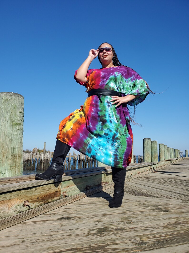 May include: A woman wearing a colorful tie-dye maxi dress with a black belt and black boots. She is standing on a wooden dock with a blue sky in the background.