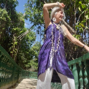 May include: A woman wearing a purple and white tie-dye top with a white shell necklace and white pants. She is standing on a path with green railings and lush greenery in the background.