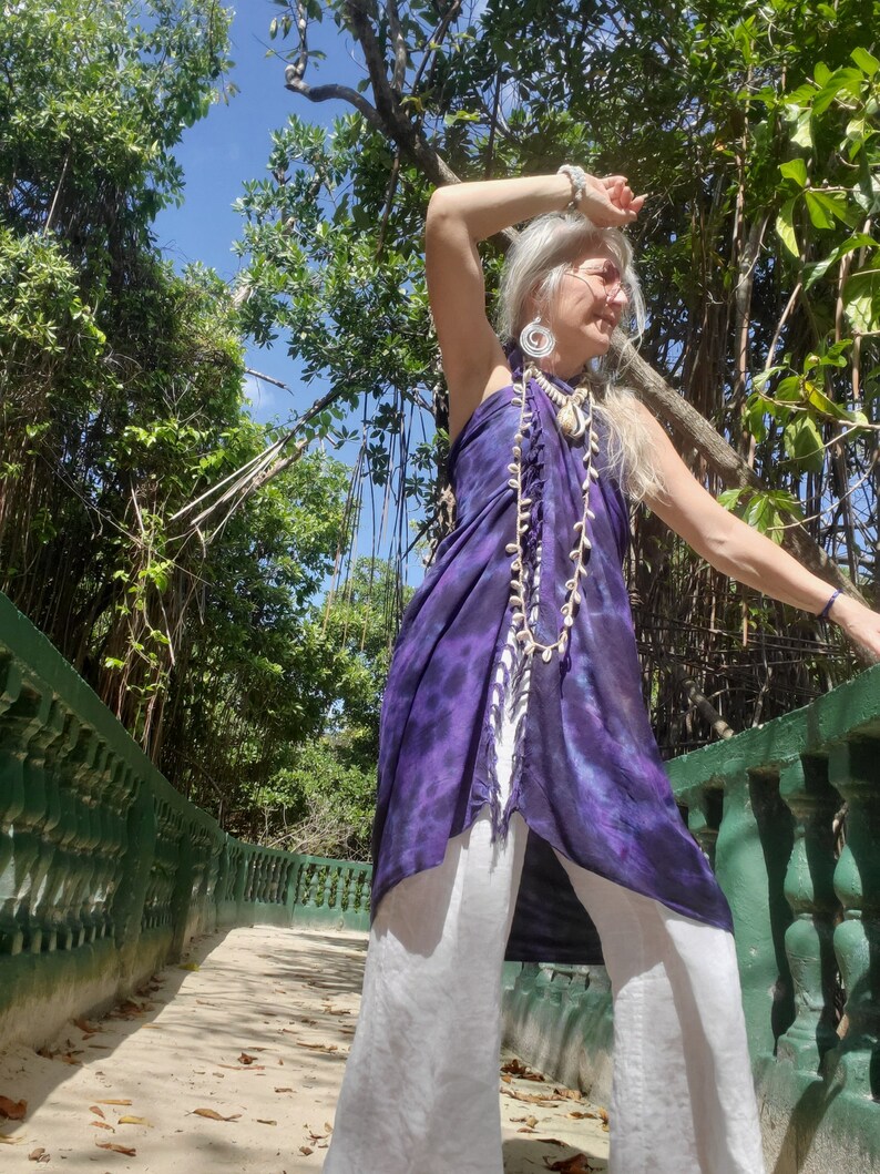 May include: A woman wearing a purple and white tie-dye top with a white shell necklace and white pants. She is standing on a path with green railings and lush greenery in the background.
