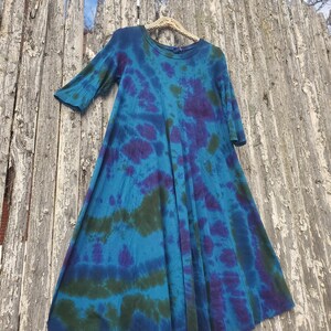 Puede incluir: Un vestido te&ntilde;ido anudado en tonos azules, morados y verdes cuelga de una valla de madera. El vestido tiene un cuello redondo y mangas tres cuartos. La tela es suave y fluida, con un patr&oacute;n &uacute;nico.