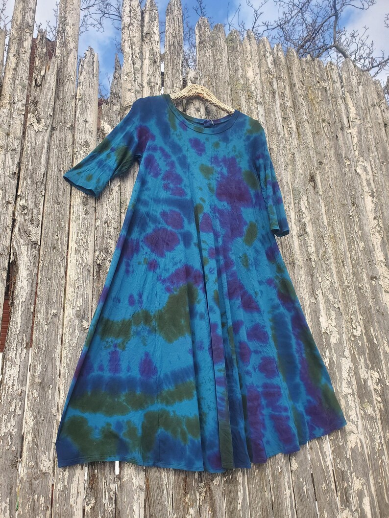 Puede incluir: Un vestido te&ntilde;ido anudado en tonos azules, morados y verdes cuelga de una valla de madera. El vestido tiene un cuello redondo y mangas tres cuartos. La tela es suave y fluida, con un patr&oacute;n &uacute;nico.