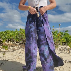 Puede incluir: Pantalones de pernera ancha con estampado tie-dye en tonos morados y negros. Los pantalones están hechos de una tela fluida y se usan en una playa de arena. La modelo lleva un top blanco y un collar de conchas. Los pantalones tienen un corte holgado.
