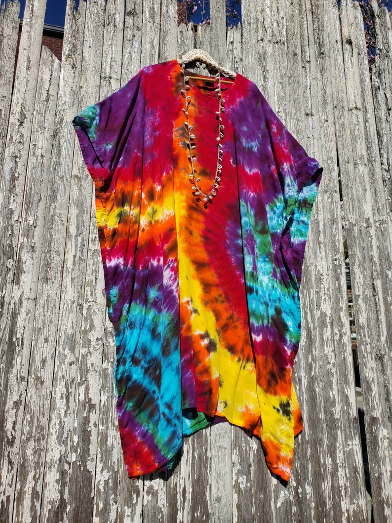 May include: A colorful tie-dye poncho with a rainbow pattern in shades of purple, red, orange, yellow, blue, and green. The poncho is hanging on a wooden fence.