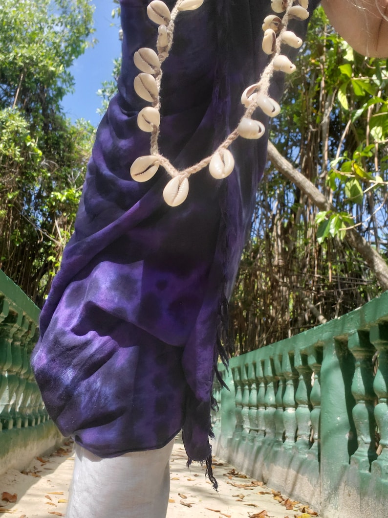 May include: A purple tie-dye sarong with a shell necklace made of cowrie shells. The sarong is worn by a person walking on a path with a green railing.