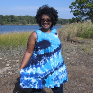 May include: A sleeveless, tie-dye tunic in shades of blue and white. The tunic has a flared silhouette and is paired with dark blue jeans and brown sandals. The background features a body of water and a clear blue sky.