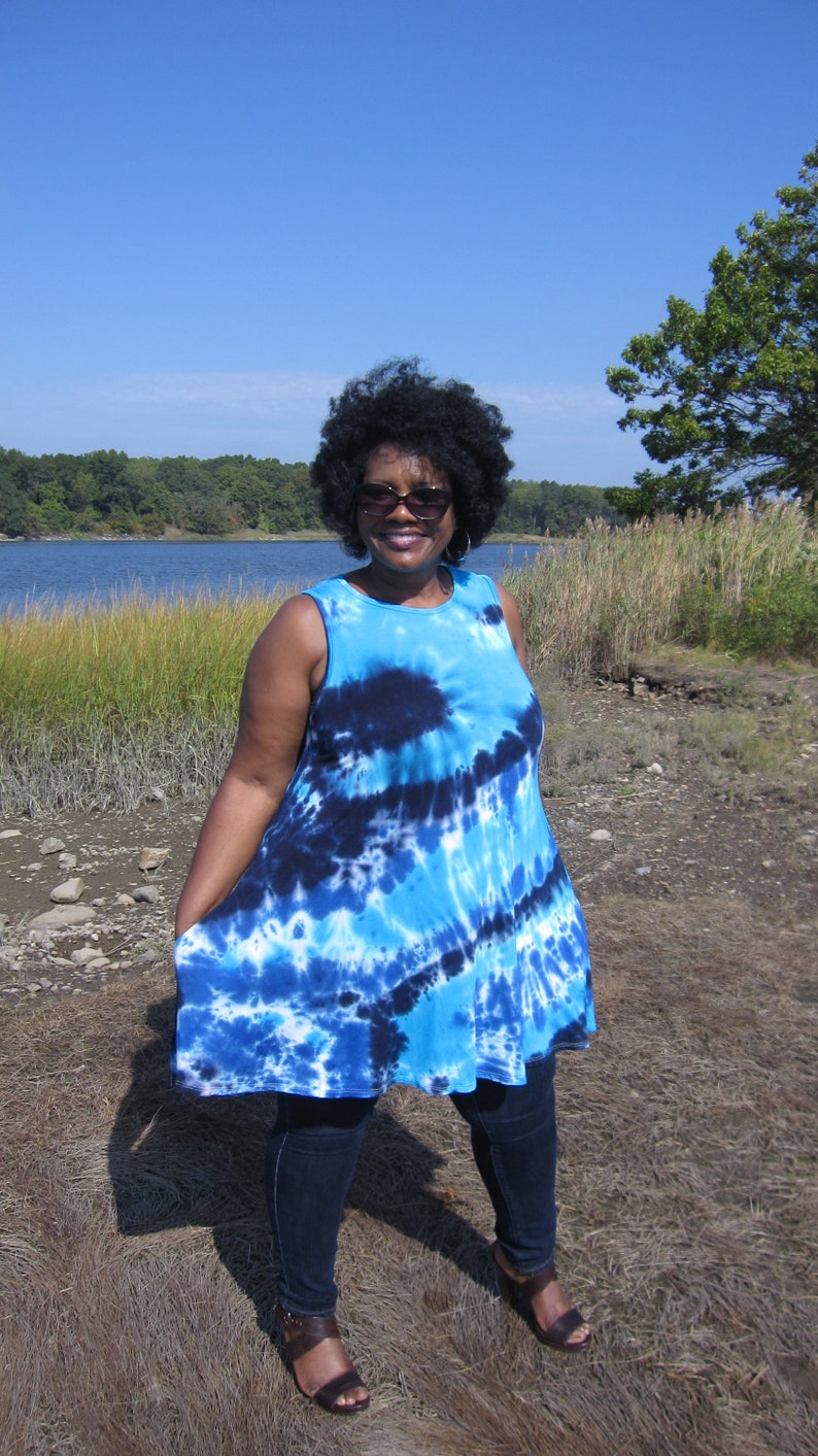 May include: A sleeveless, tie-dye tunic in shades of blue and white. The tunic has a flared silhouette and is paired with dark blue jeans and brown sandals. The background features a body of water and a clear blue sky.