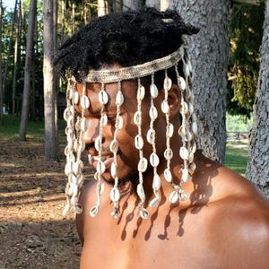 Può includere: Copricapo decorativo con una fascia intrecciata ornata da fili pendenti di conchiglie bianche. L'accessorio è indossato su uno sfondo di alberi, con ombre create dalla luce solare.