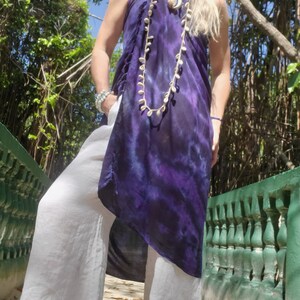 May include: A woman wearing a long, purple and blue tie-dye dress with a white shell necklace and white pants.